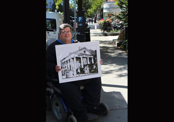 Allen Chamberland poses with one of his paper cuts depicting Boston’s Quincy Market. Chamberland, who suffers from a chronic condition that affects his breathing, hopes the success he has selling his artwork on ArtLifting will help him get off Social Security. Photo by Liz Powers