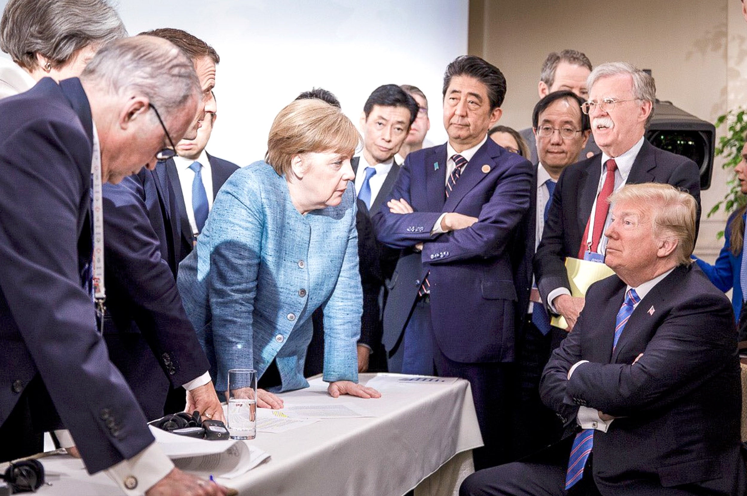 Angela Merkel, surrounded by world leaders at G& summit in 2018, leans over table to face Donald Trump, whose arms are folded.