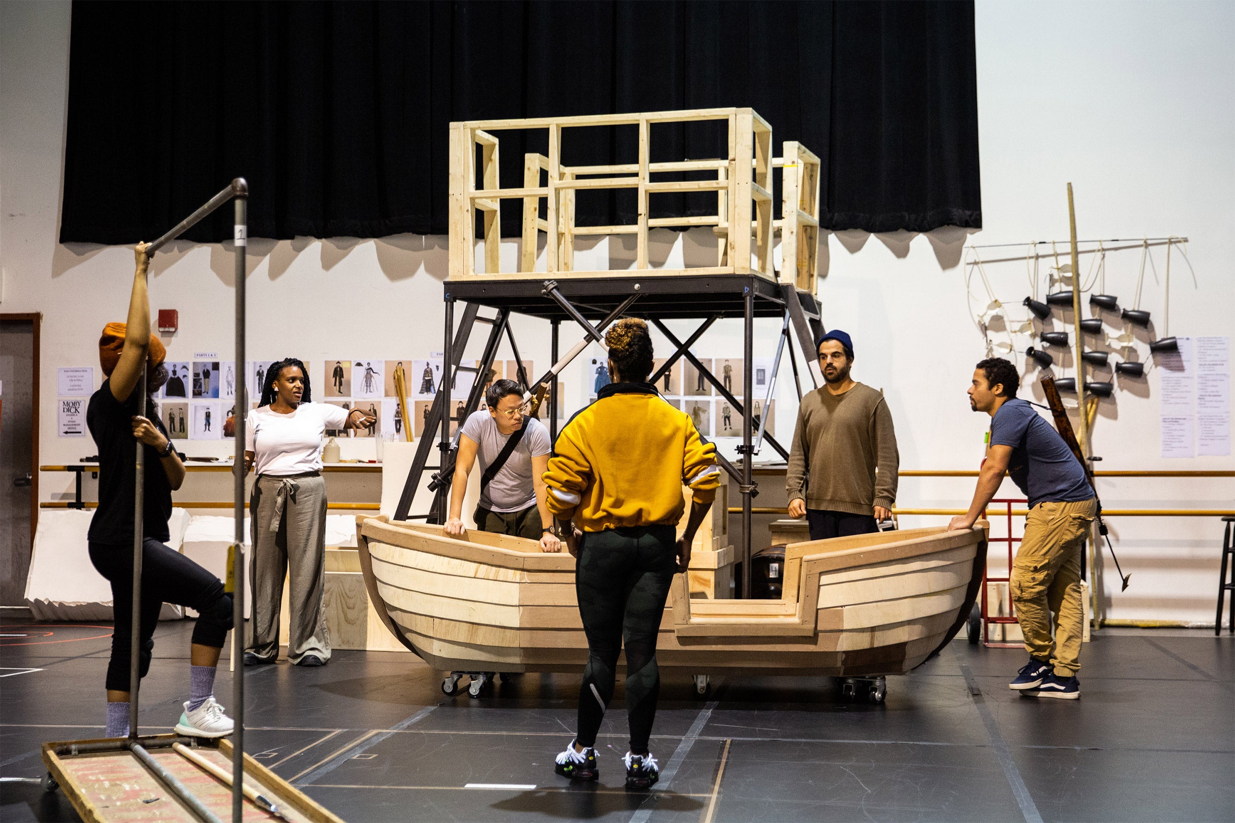 Cast members stand around a wooden prop boat as a choreographer looks on in the center.
