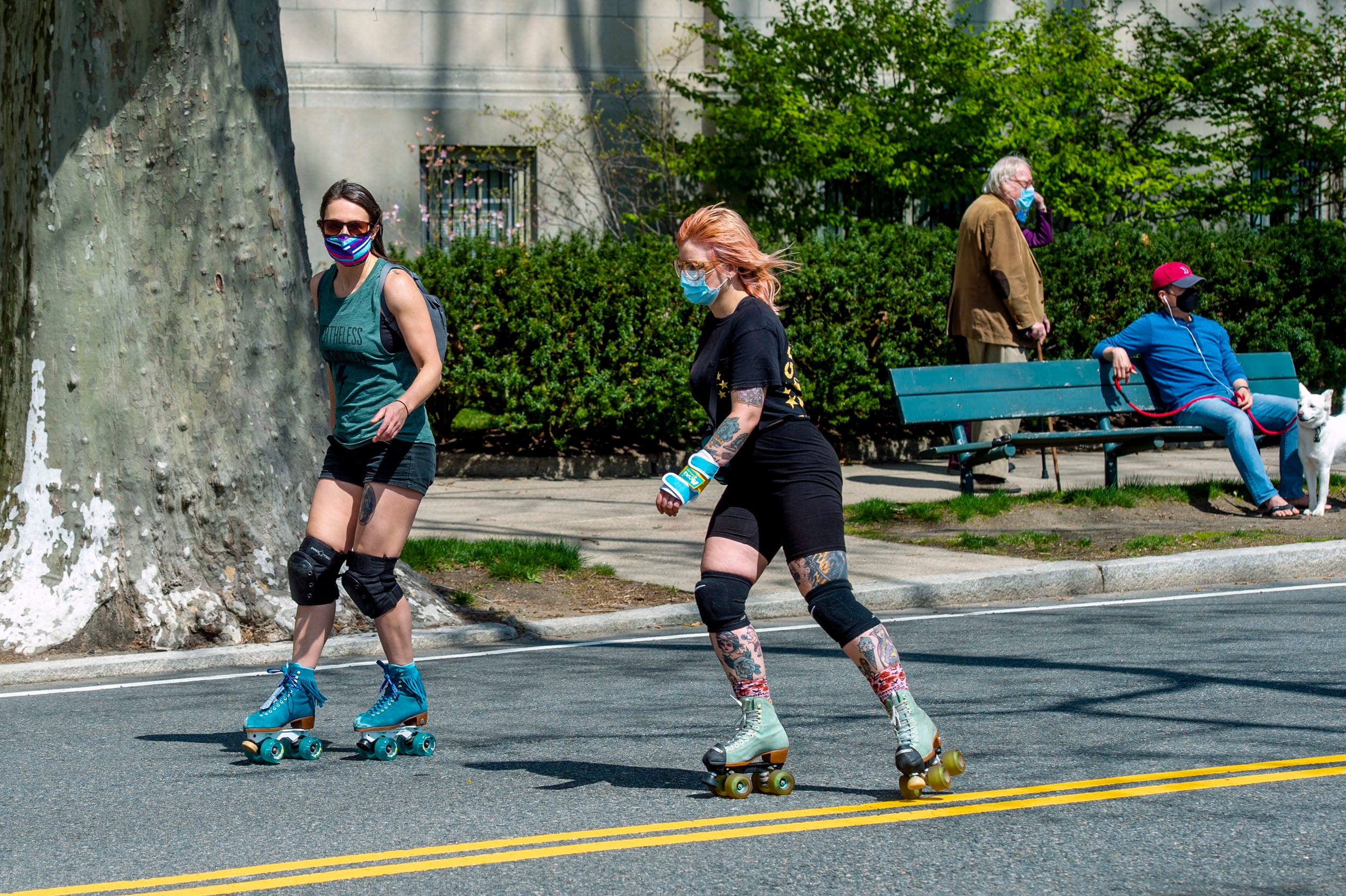 A couple of Rollerbladers glide past a walker and a man sitting on a bench with his dog.