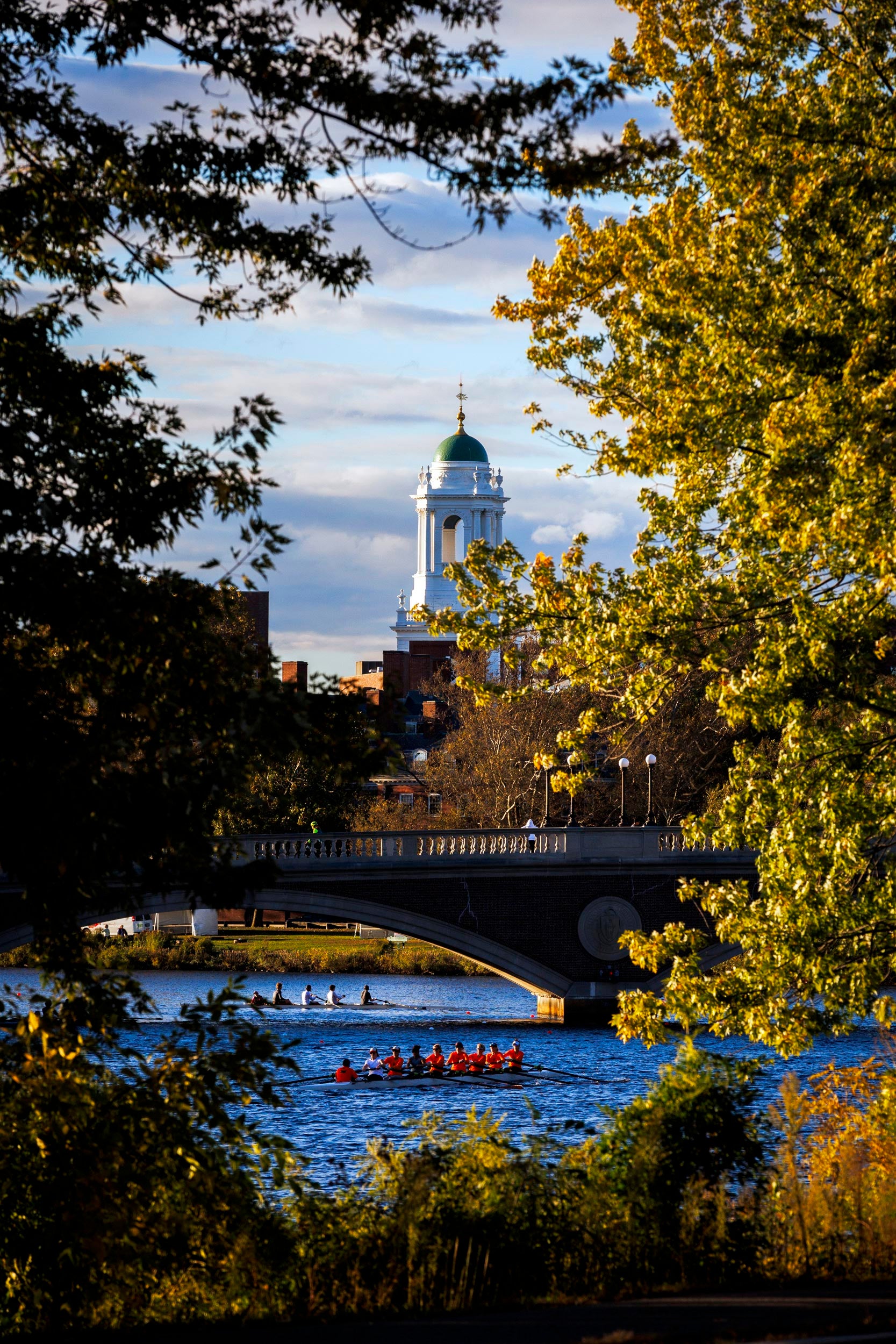 Rowers are pictured along the Charles River with the Eliot House tower in the background.