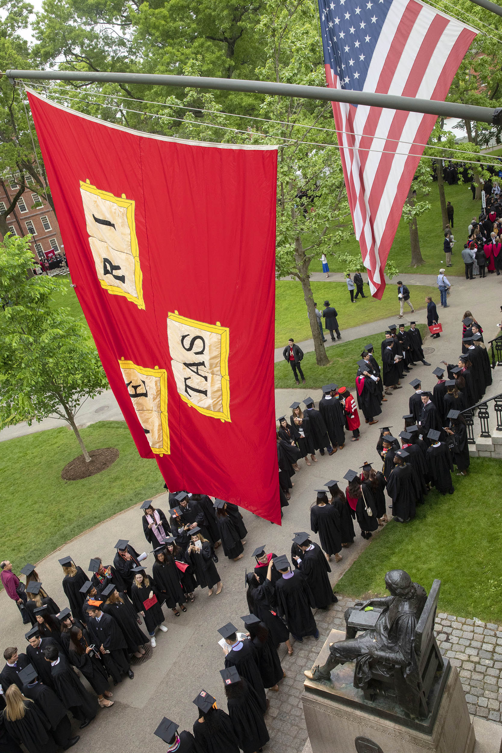 During last year’s Morning Exercises at Harvard’s 368th Commencement, graduates join the procession beneath veritas flags and the John Harvard Statue in the Tercentenary Theatre.