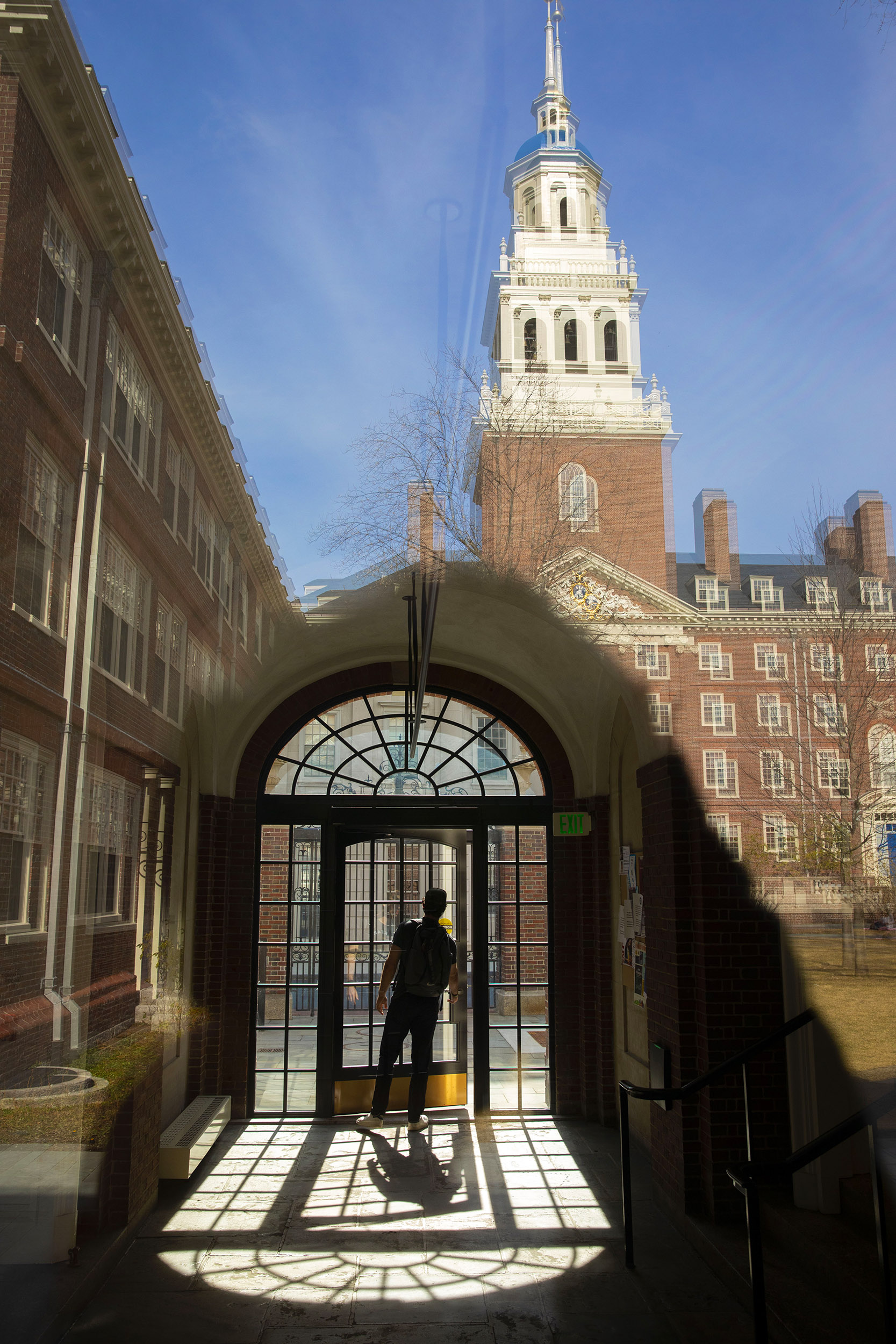 A student is framed by a bay window inside the passageway leading to the dining hall in Lowell House courtyard.