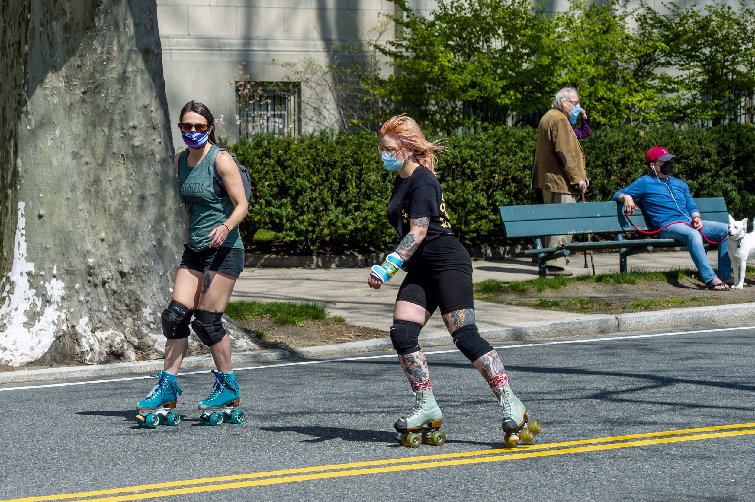 A couple of Rollerbladers glide past a walker and a man sitting on a bench with his dog.