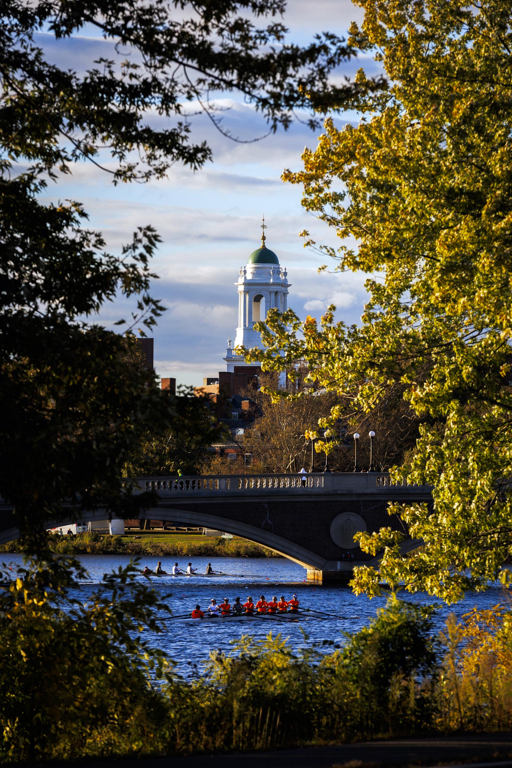 Rowers are pictured along the Charles River with the Eliot House tower in the background.