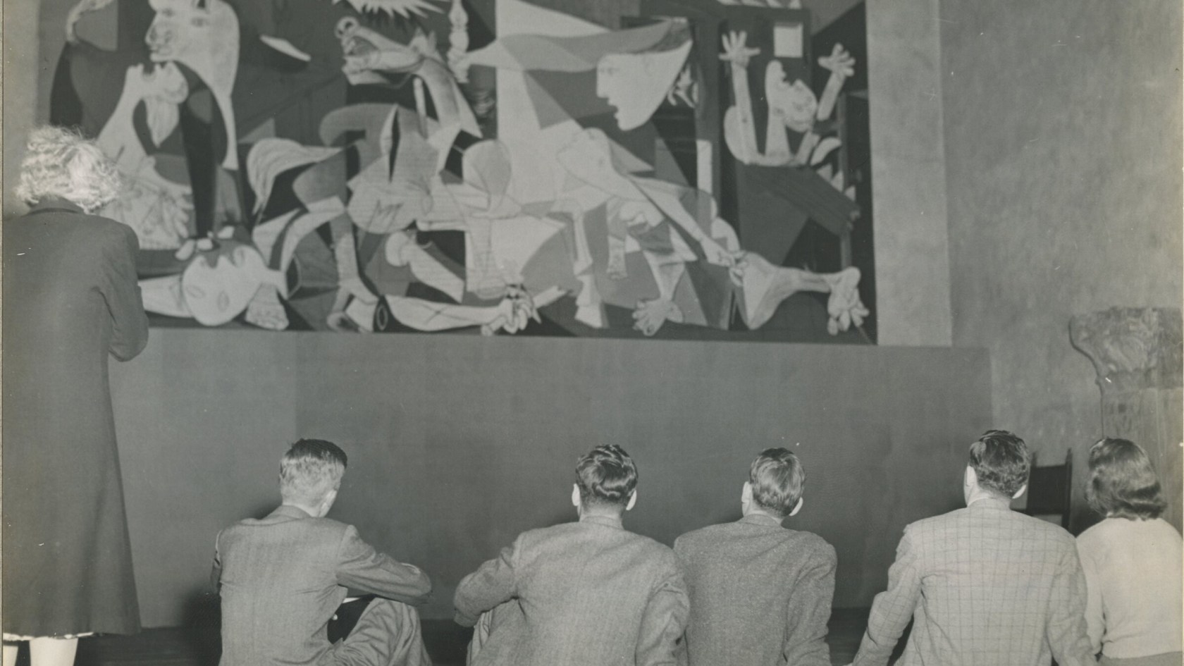 One woman standing and four men sitting in from of the oil painting Guernica by Pablo Picasso. 
