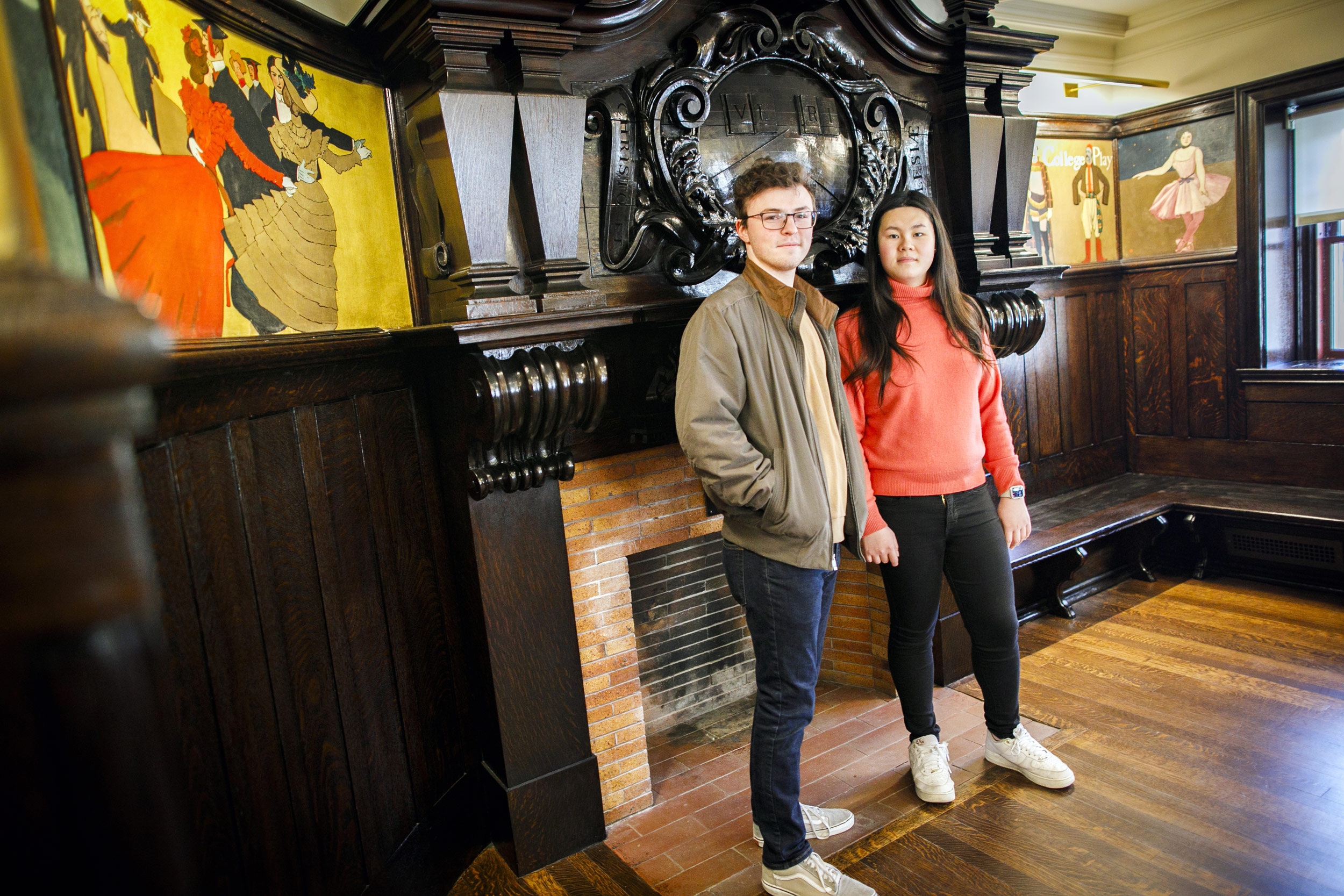 Adams House’s House Committee Chairs (aka HoCos) Lily Liu and Tate Underwood share their favorite aspects of their student residence. Tate Underwood (left) and Lily Liu are pictured in the Coolidge Room by a fireplace decorated with a wooden carved veritas shield at Adams House.