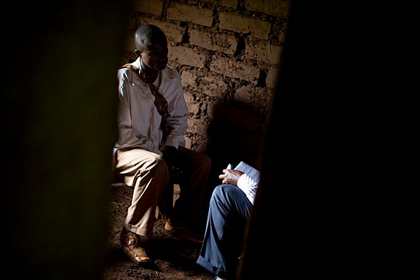 Two men sit close, knees almost touching, in a mud-walled hut in the Congolese village of Katokota.