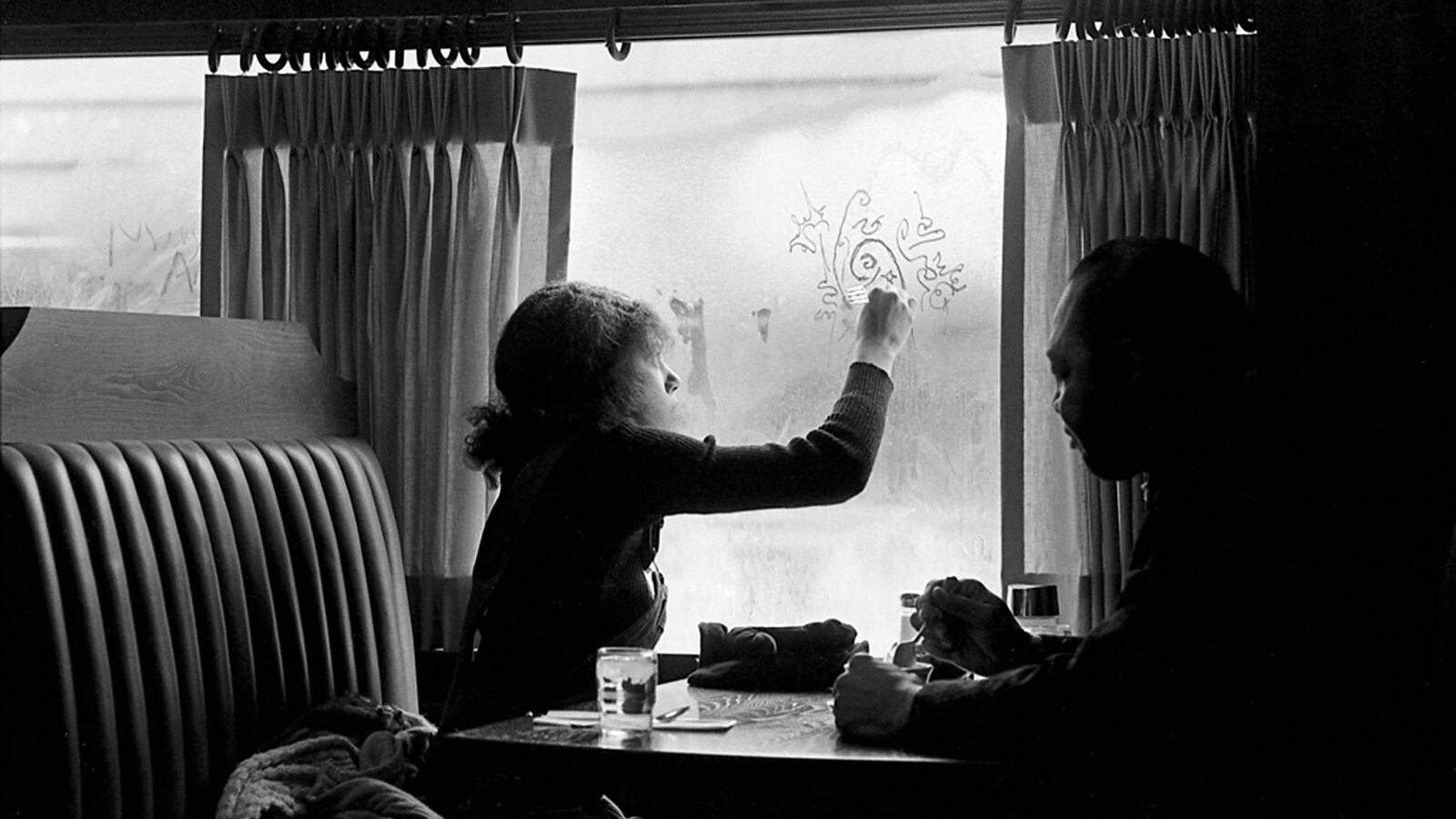 Woman in restaurant booth doodles in window fog, 1969.
