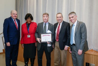 Five people posing for a photo; man in the middle holds an award certificate.