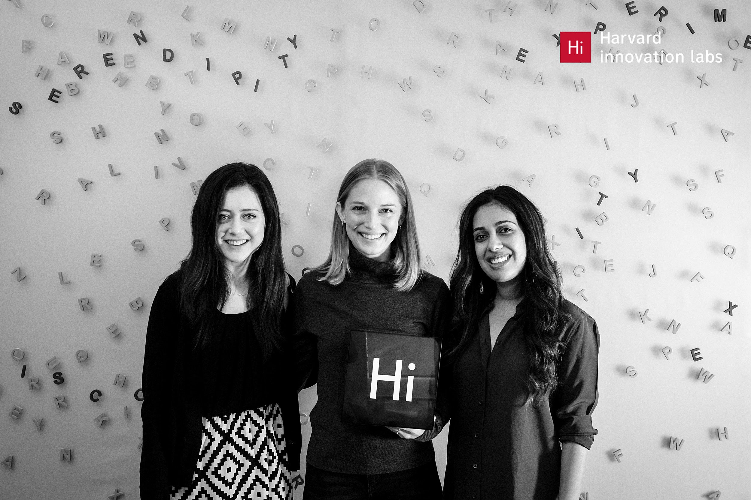 Three women standing together holding a small sign that reads "Hi"