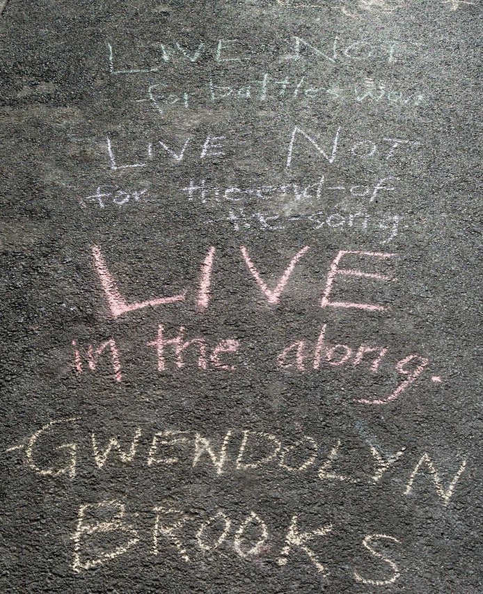 sidewalk chalk on the street that says “Live not for Battles Won. Live not for The-End-of-the-Song. Live in the along. Gwendolyn Brooks"