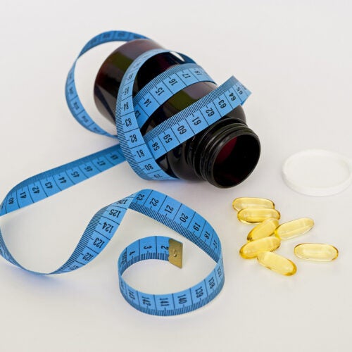 A blue measuring tape wrapped around a medicine bottle, with loose pills scattered around the open lid.