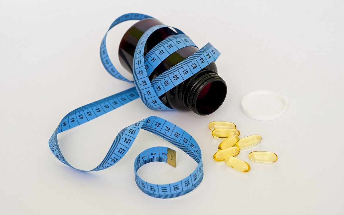 A blue measuring tape wrapped around a medicine bottle, with loose pills scattered around the open lid.