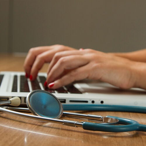 Typing on a laptop with stethoscope on desk.