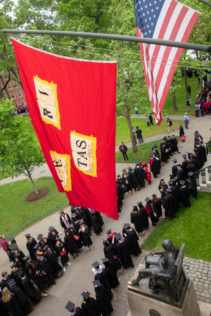 During last year’s Morning Exercises at Harvard’s 368th Commencement, graduates join the procession beneath veritas flags and the John Harvard Statue in the Tercentenary Theatre.
