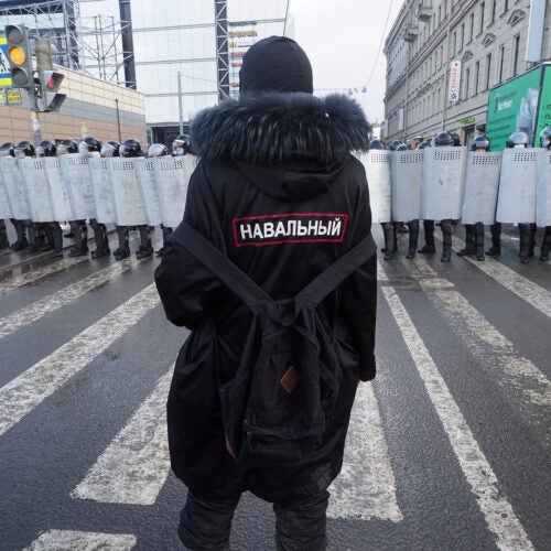 A man with a sign 'Navalny' on his back stands in front of riot policeme.