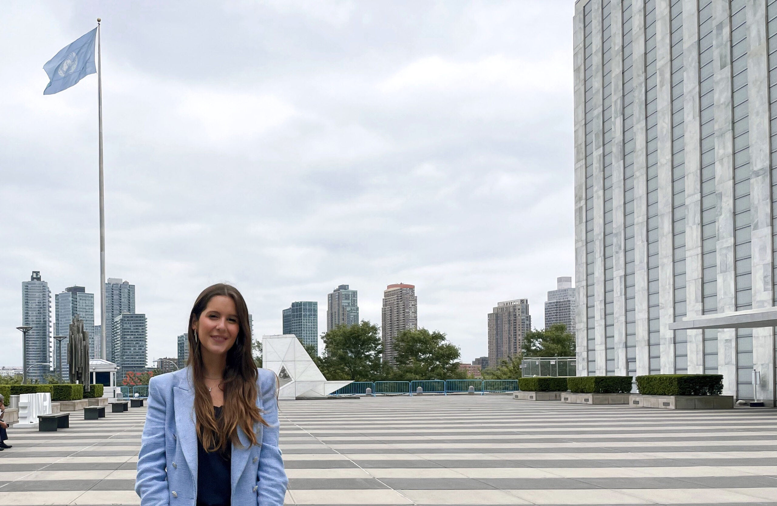 View of a young woman in a light blue jacket standing at United Nations Plaza and building entrance with the United Nations' flag visible.