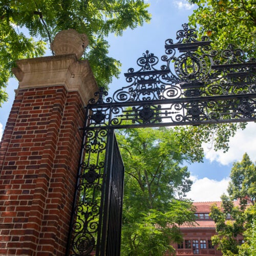 Sever Hall and a gate with an “H” is framed by foliage in Harvard Yard.