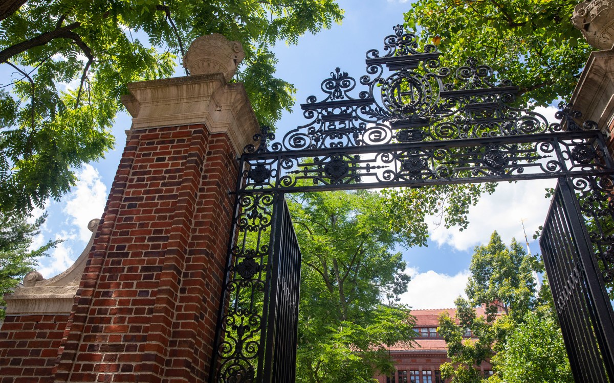 Sever Hall and a gate with an “H” is framed by foliage in Harvard Yard.