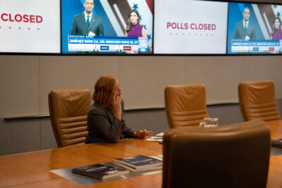 In scene from "Succession," Shiv Roy talks in the phone in an ATN board room with election coverage on TVs in background.