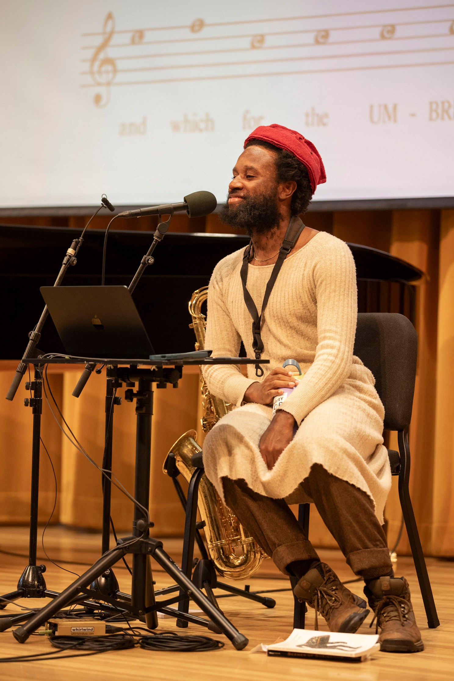 Jerome Ellis sits and speaks into microphone on Paine Hall stage with piano, saxophone, and laptop. A screen behind him projects poetry and sheet music.