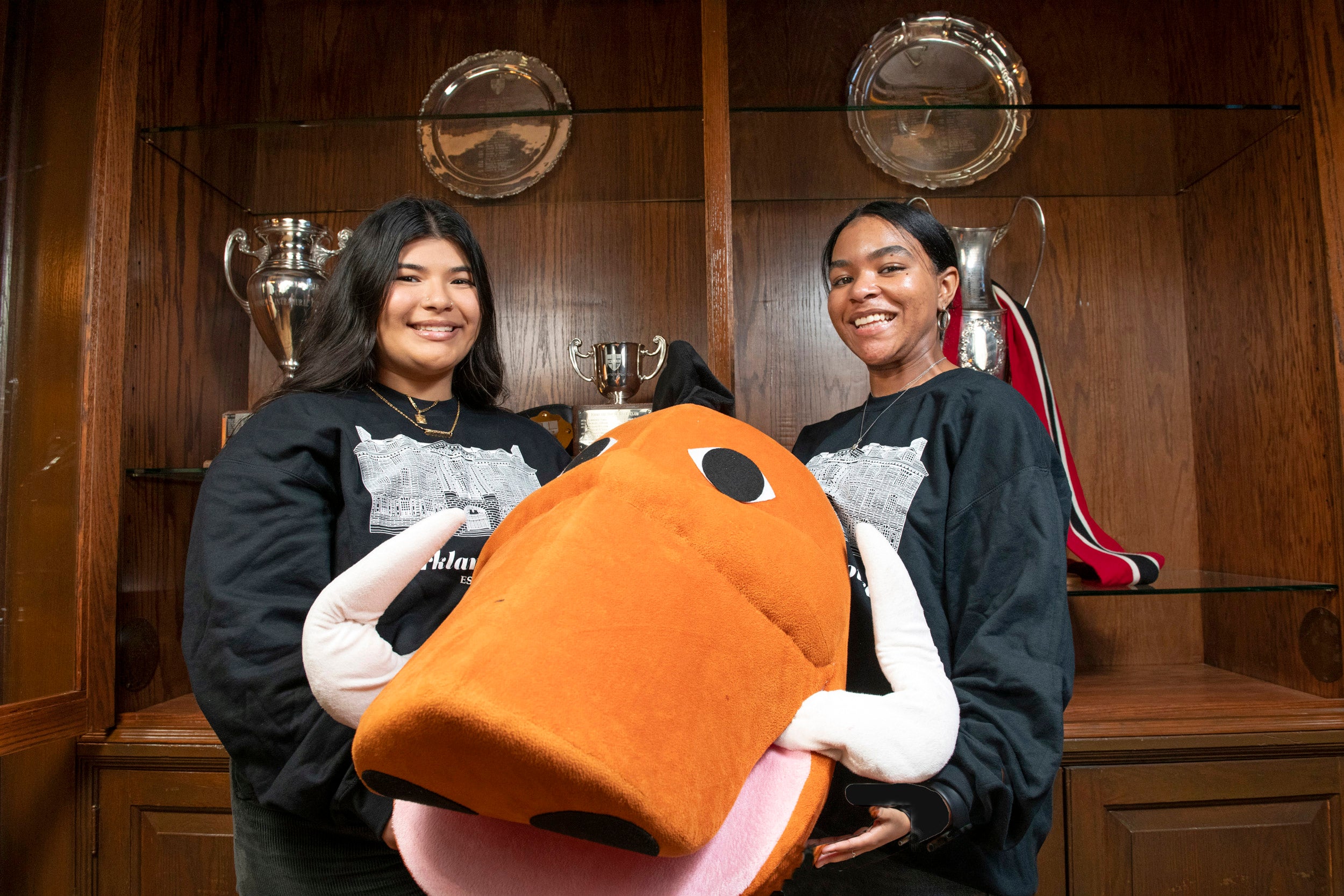 Giselle Chiprez (left) and Nena King with their boar mascot costume in front of a trophy case in the Kirkland House Junior Common Room.