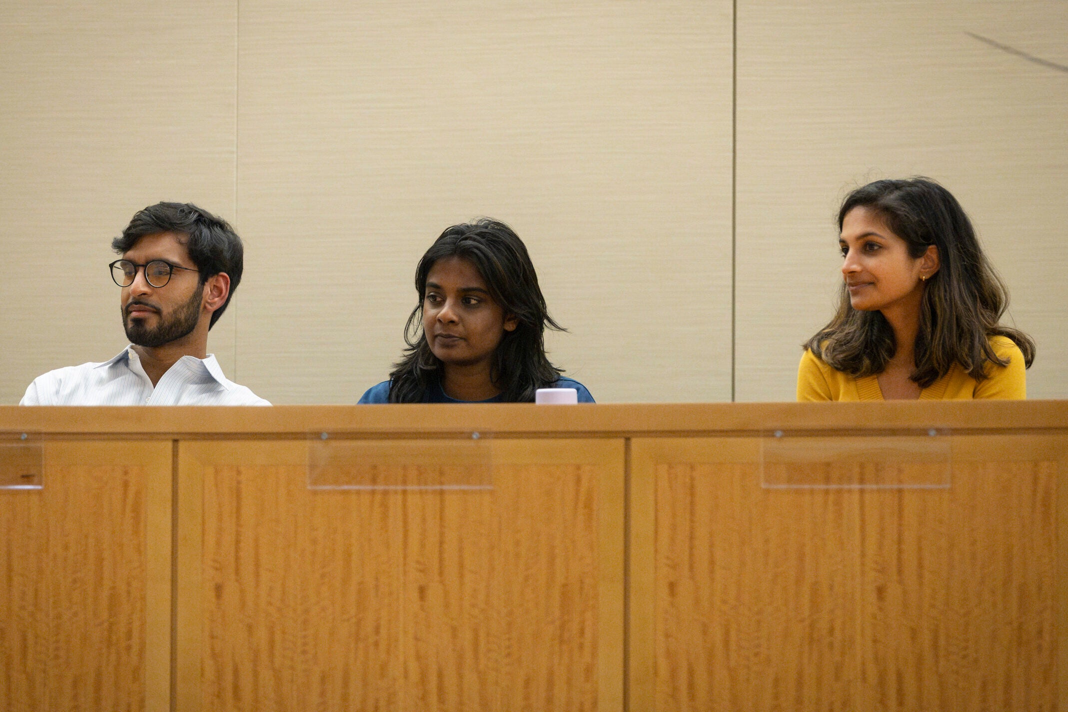 Three students listening to the speakers while sitting behind a wooden desk.
