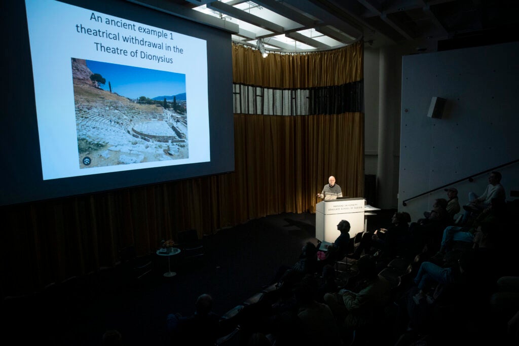 Richard Sennett speaking at a podium while showing a slide of the theater of dionysus. 