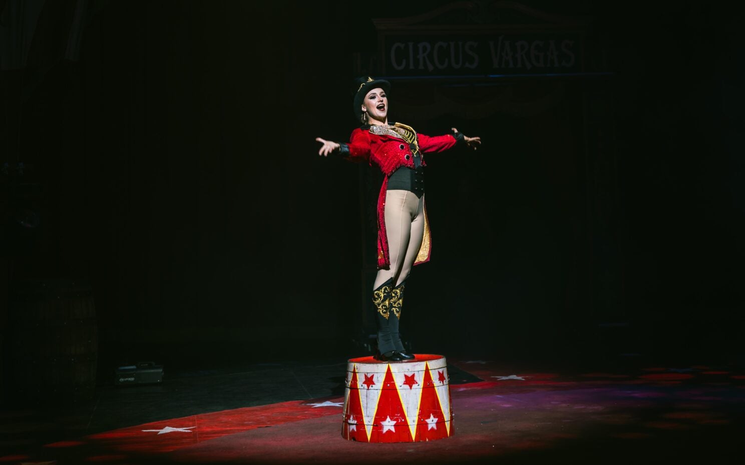 Performer Izzy Patrowicz wearing costume including top hat stands on a barrel at the circus.