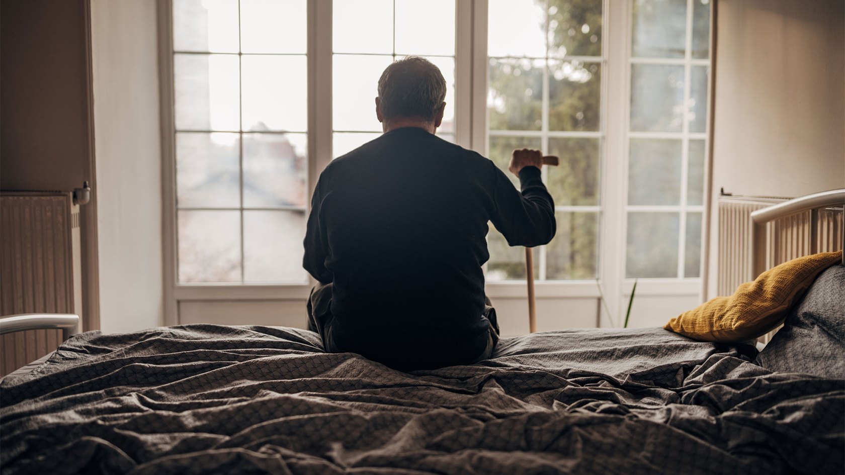 A lonely elderly man is sitting on the bedlooking out the window.