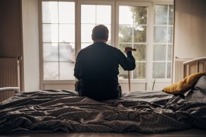 A lonely elderly man is sitting on the bedlooking out the window.