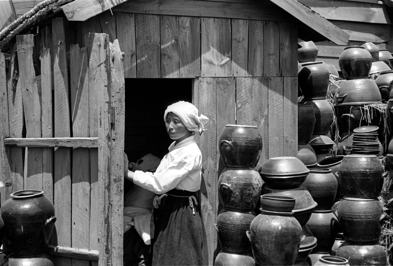 A woman stands in the doorway of a wooden building, framed by stacks of jars