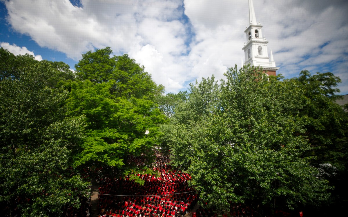 Graduates are seen through the windows of Sever Hall sitting in Tercentenary Theatre with the steeple of Memorial Church rising into the sky.