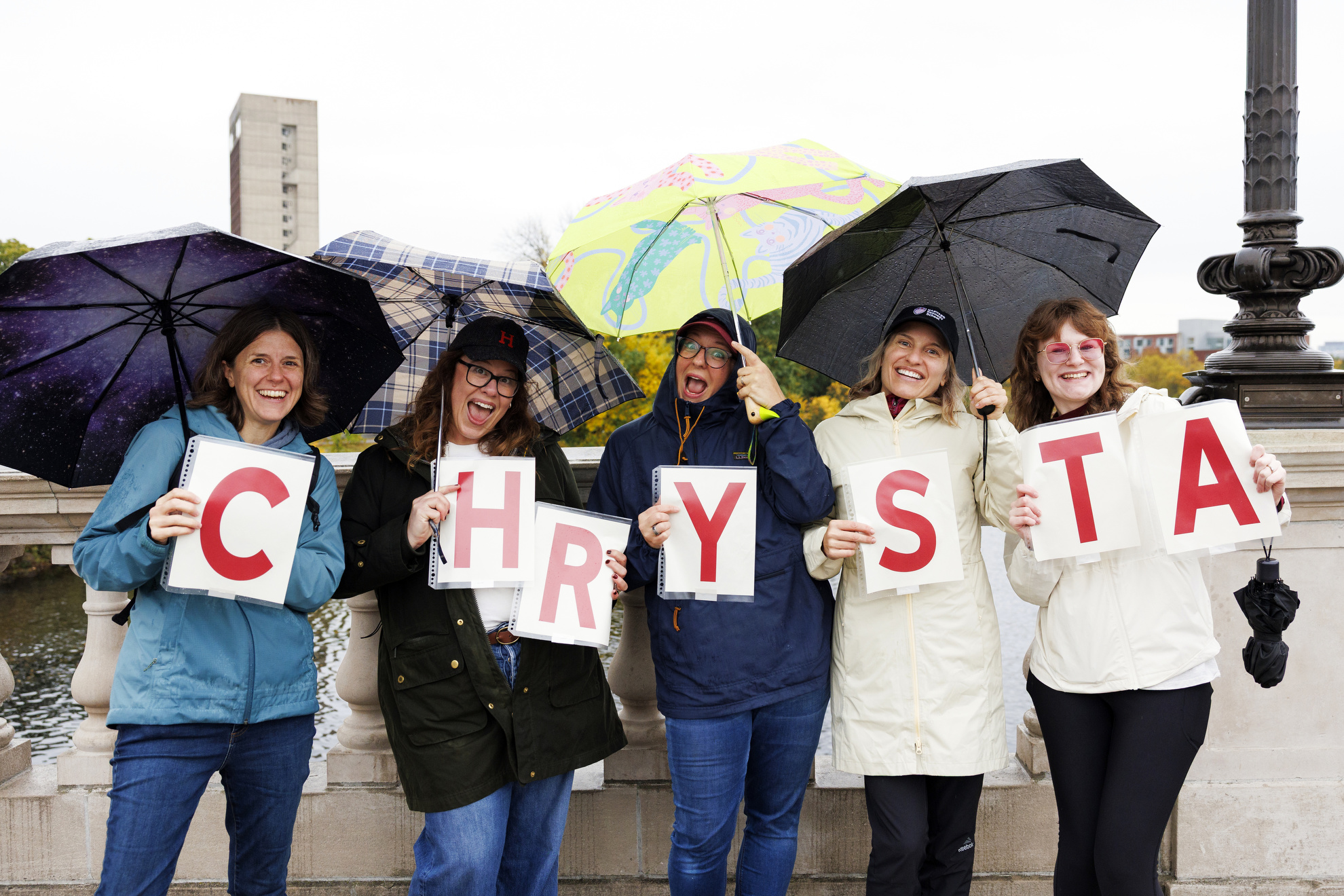 Harvard Kennedy School MC/MPA students pose for a photo on the John W. Weeks Footbridge with a sign they made to cheer on their classmate, Chrysta Castanñeda MC/MPA ’27, who is competing.