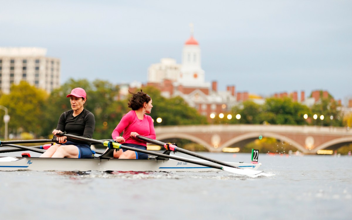 Two rowers head towards the John W. Weeks Footbridge with Dunster House visible in the background.
